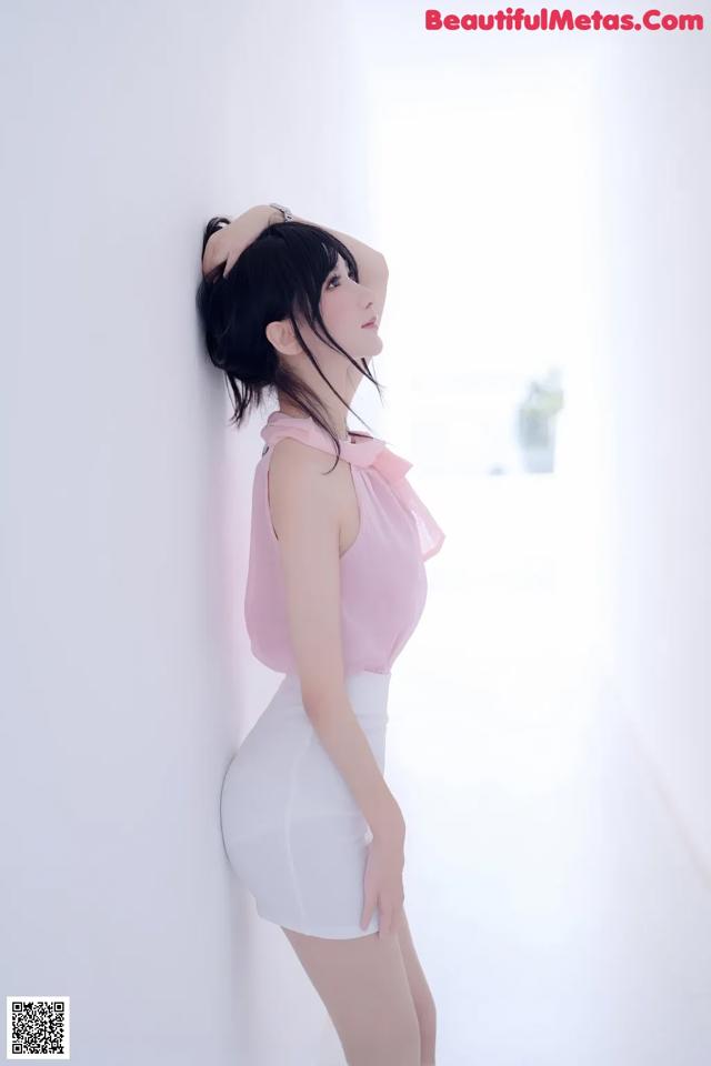 A woman in a pink top and white skirt leaning against a wall.